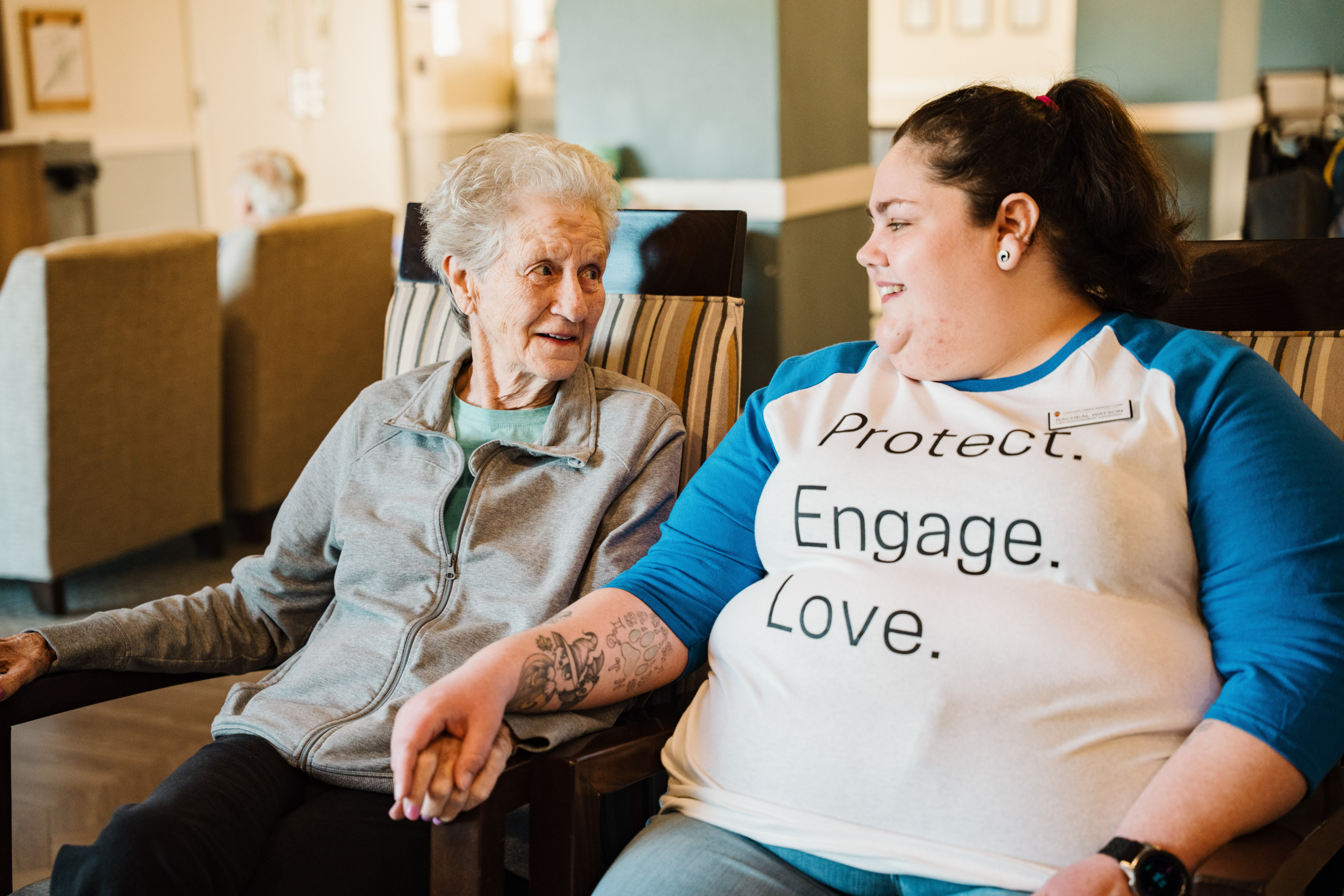 A resident and staff member are sitting next to each other holding hands, looking at each other and smiling.